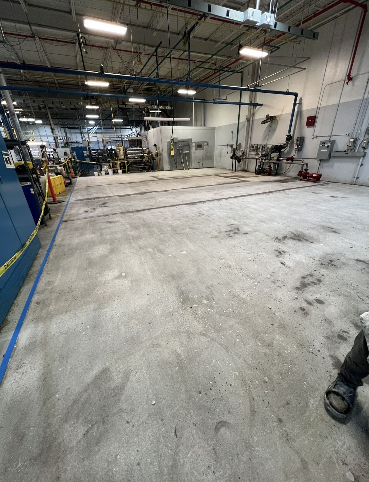 Bare, dust-covered concrete floor in a manufacturing bay before coating, with machinery and pipework along the wall